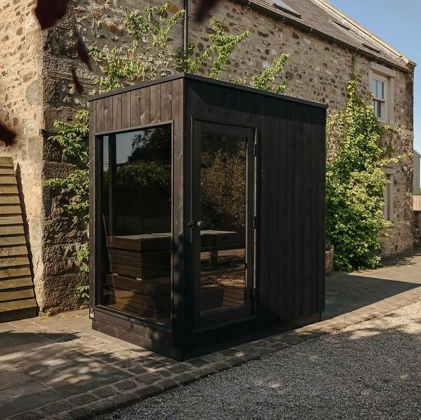 Modern black garden office pod with large glass door, beside a rustic stone cottage and leafy courtyard.