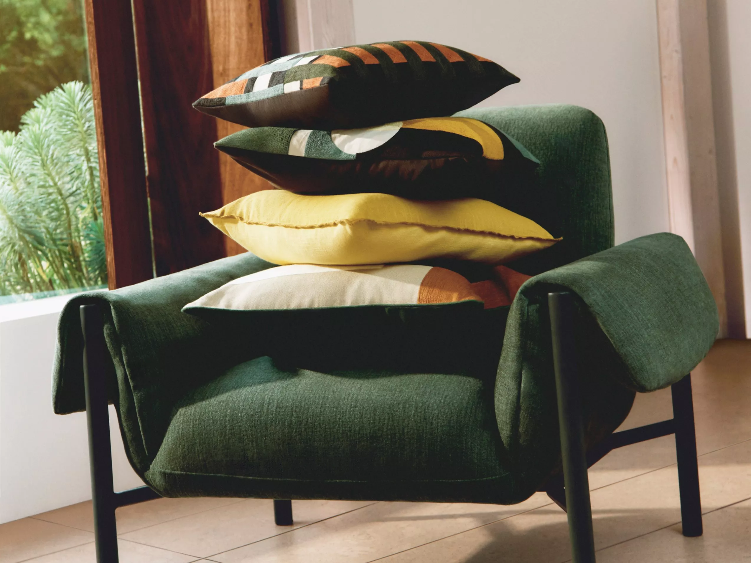 A stylish green armchair with a stack of colourful cushions, set against a window with lush greenery outside.