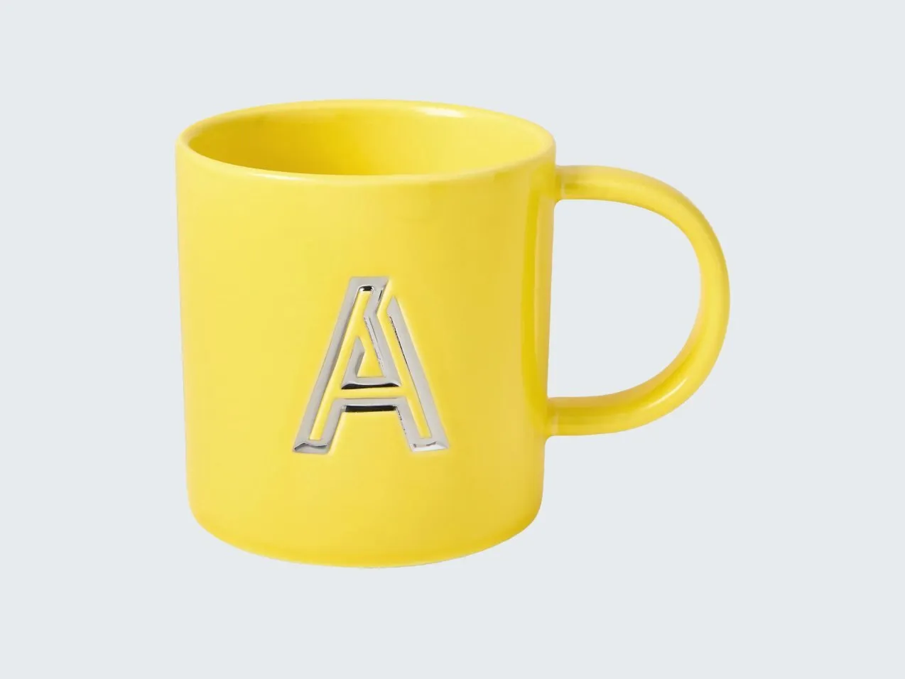 Bright yellow ceramic mug with silver letter A emblem, studio product photo on a clean light background