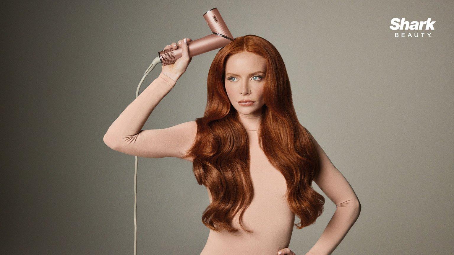 Woman with long, wavy red hair using a pink Shark Beauty hair tool against a neutral background.