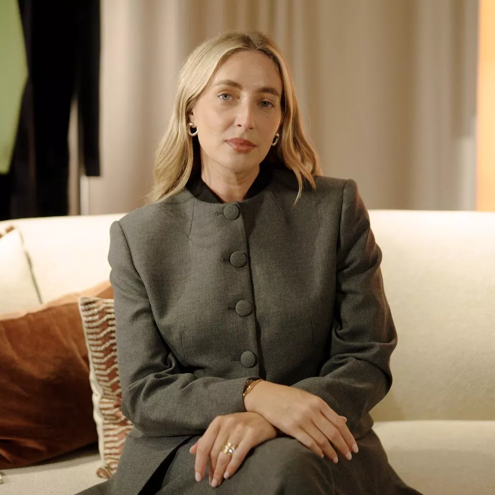 Portrait of a blonde woman in a grey tailored suit sitting on a cream sofa in a modern living room.