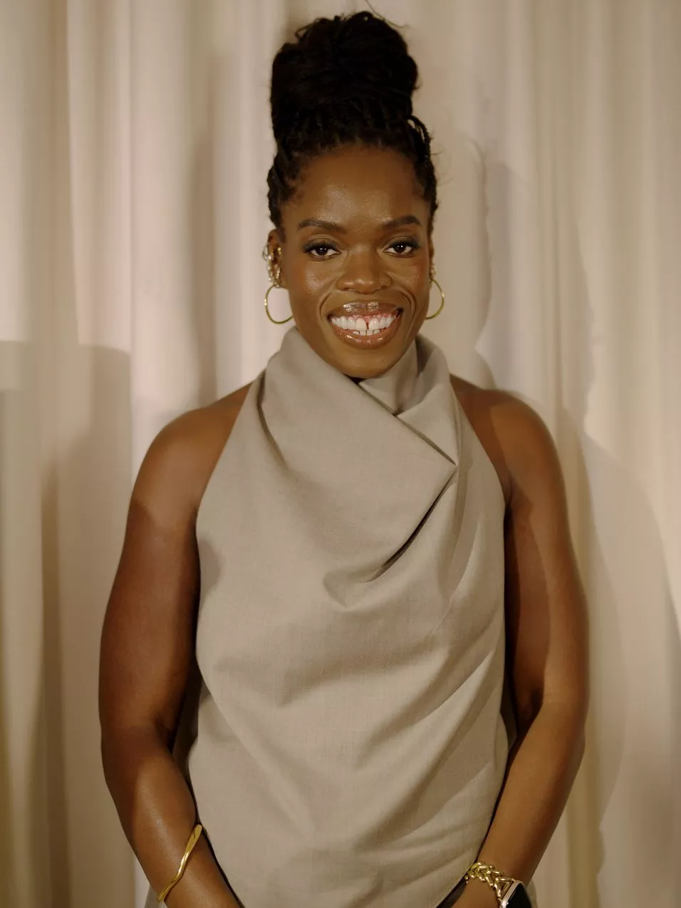 Smiling woman in beige sleeveless dress with hoop earrings, posing against cream curtain backdrop in studio portrait