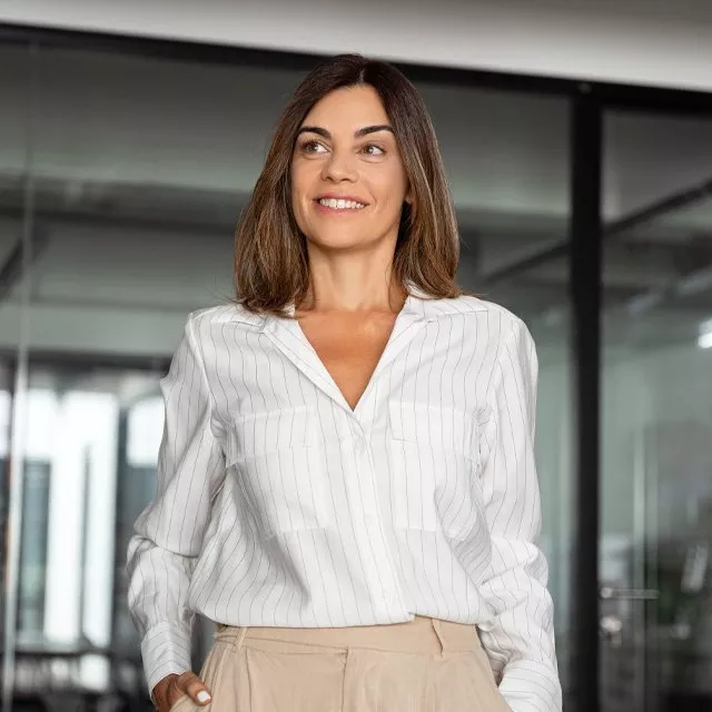 Smiling businesswoman in a modern office, wearing a white striped blouse and beige trousers, looking away