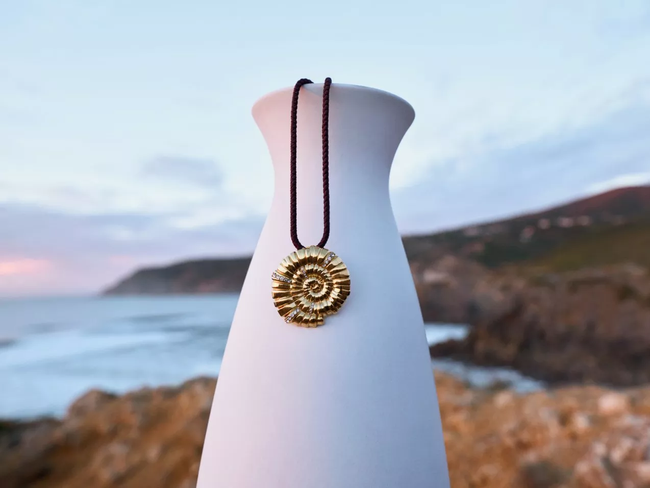 Gold ammonite pendant on a vase with a coastal backdrop, featuring cliffs and a serene sea view at dusk.