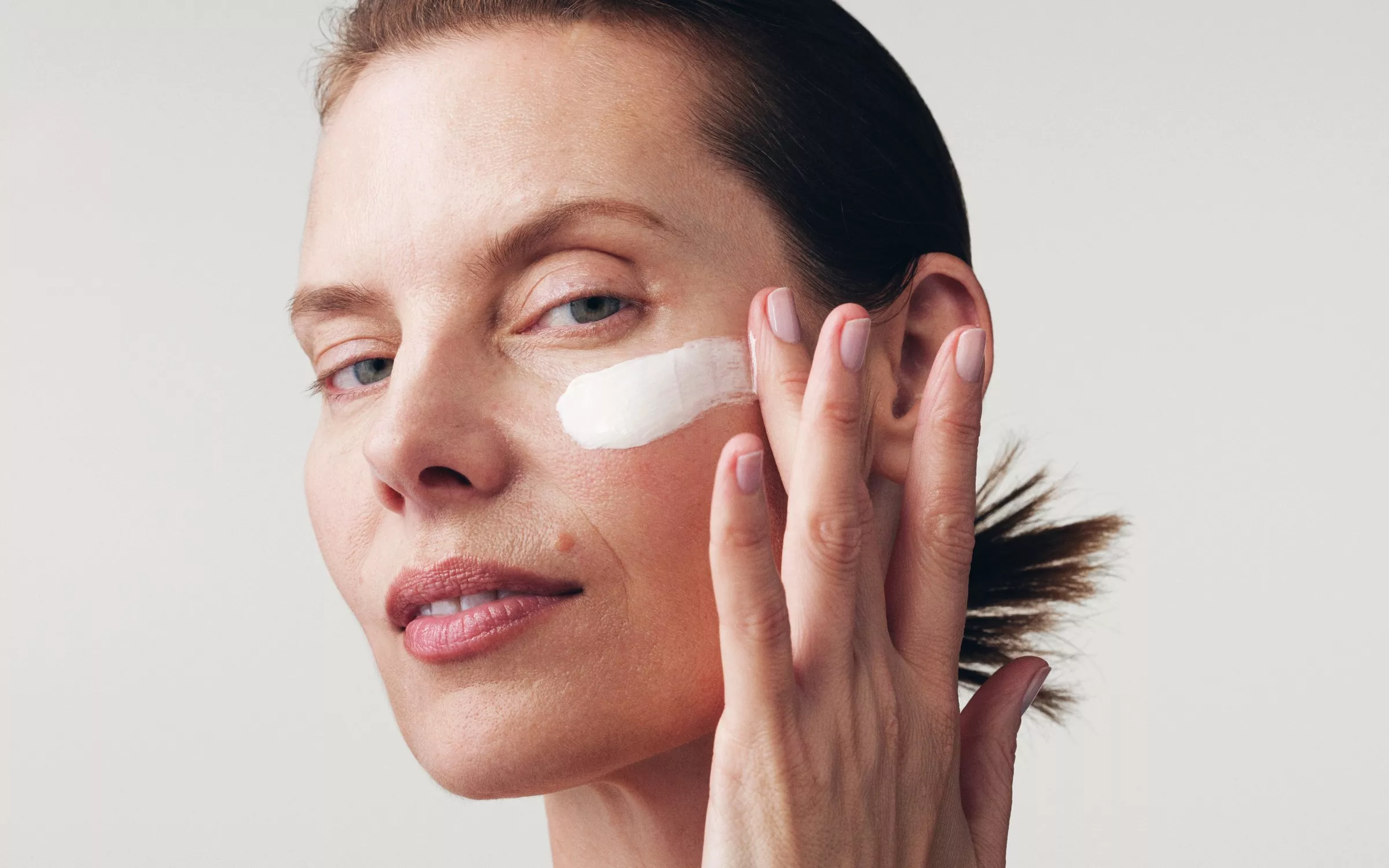  Close-up of a person with fair skin applying a white cream or moisturizer just beneath their eye with their fingertips.