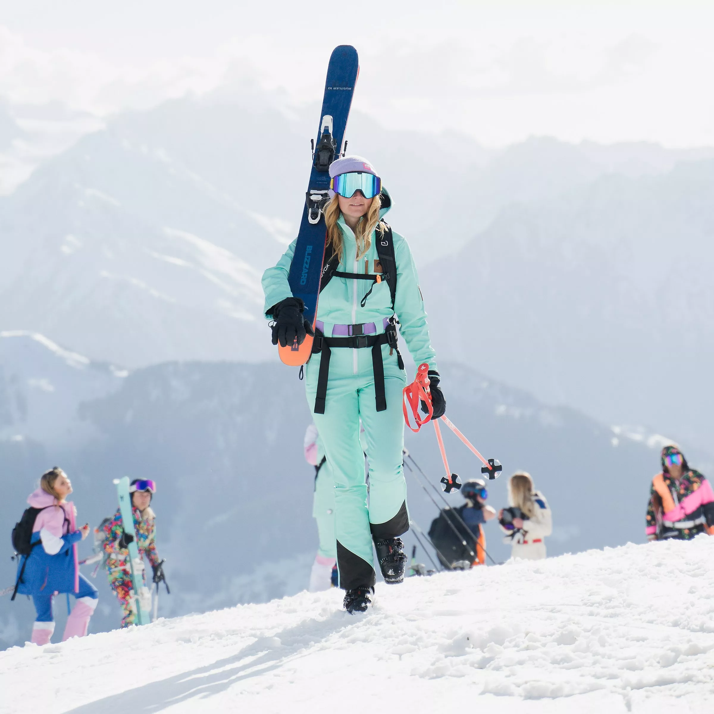 Female skier carrying skis on a snowy mountain slope, wearing goggles and a turquoise ski suit at a ski resort