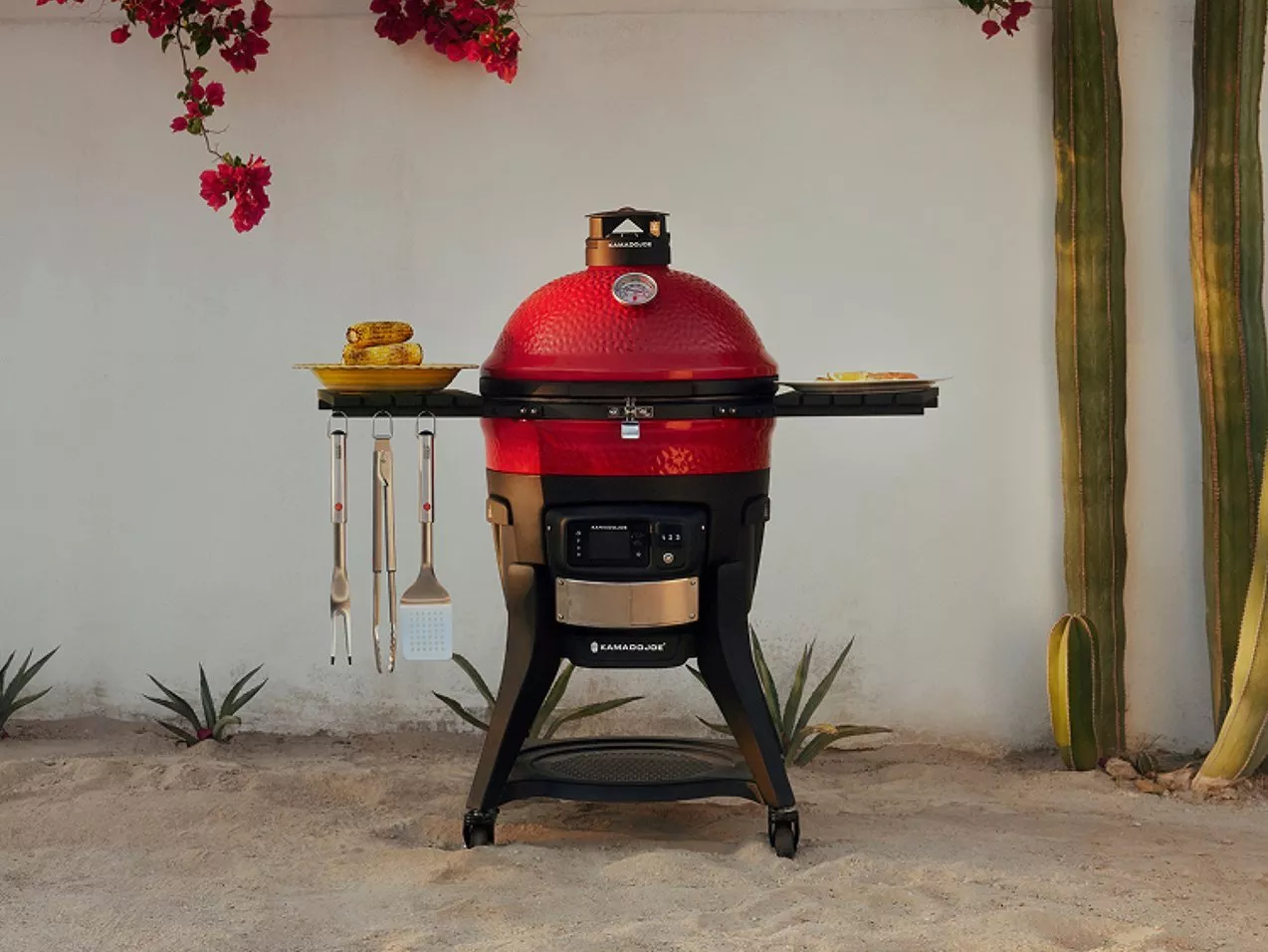 Red Kamado Joe ceramic charcoal barbecue grill with side shelves, utensils and corn, set on a sandy ground.