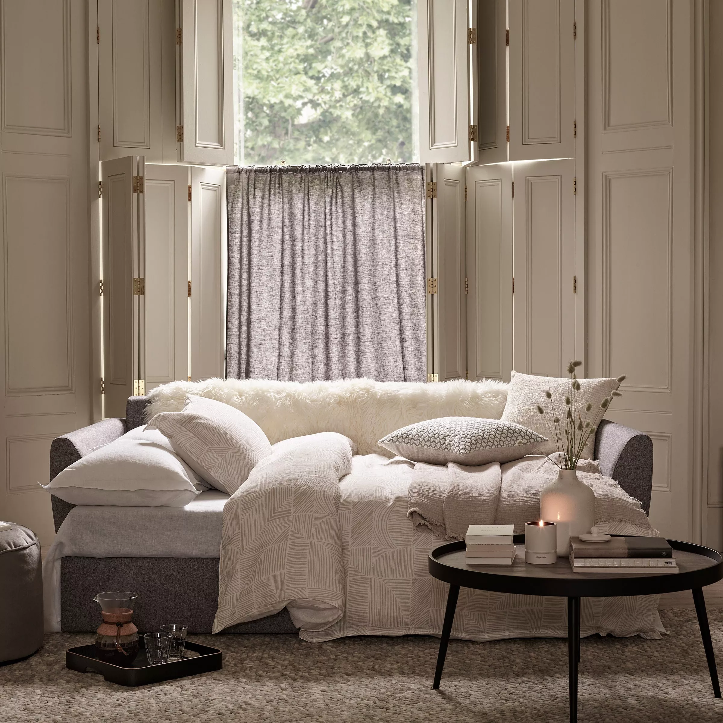 Cosy living room with grey sofa bed, neutral cushions and throws, and shuttered window with linen café curtain