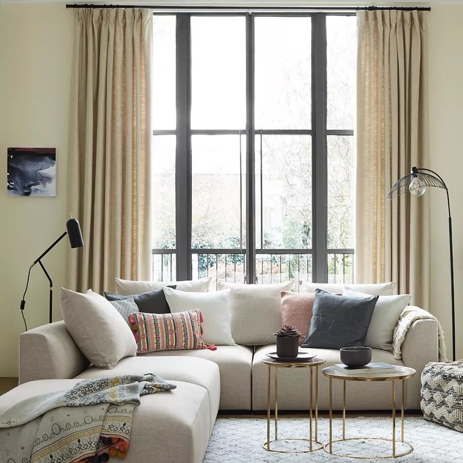 Modern living room with beige corner sofa, cushions, gold nesting tables and tall window with curtains