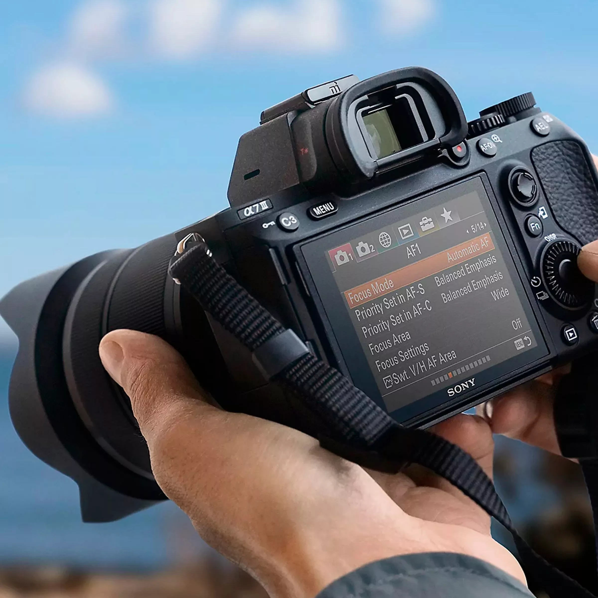 Hands holding Sony mirrorless camera showing autofocus settings on rear screen, with blue sky background outdoors