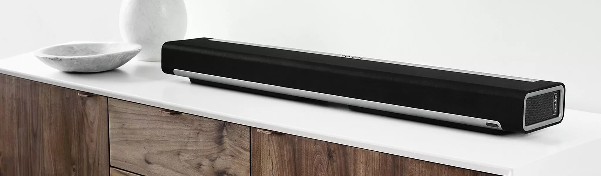 A sleek black soundbar on a white cabinet, next to a marble bowl and vase, against a minimalist background.