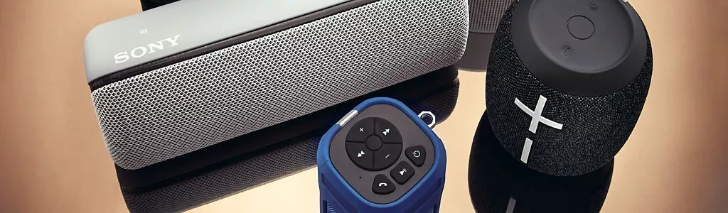 Various portable speakers in different designs and colours displayed against a reflective surface.
