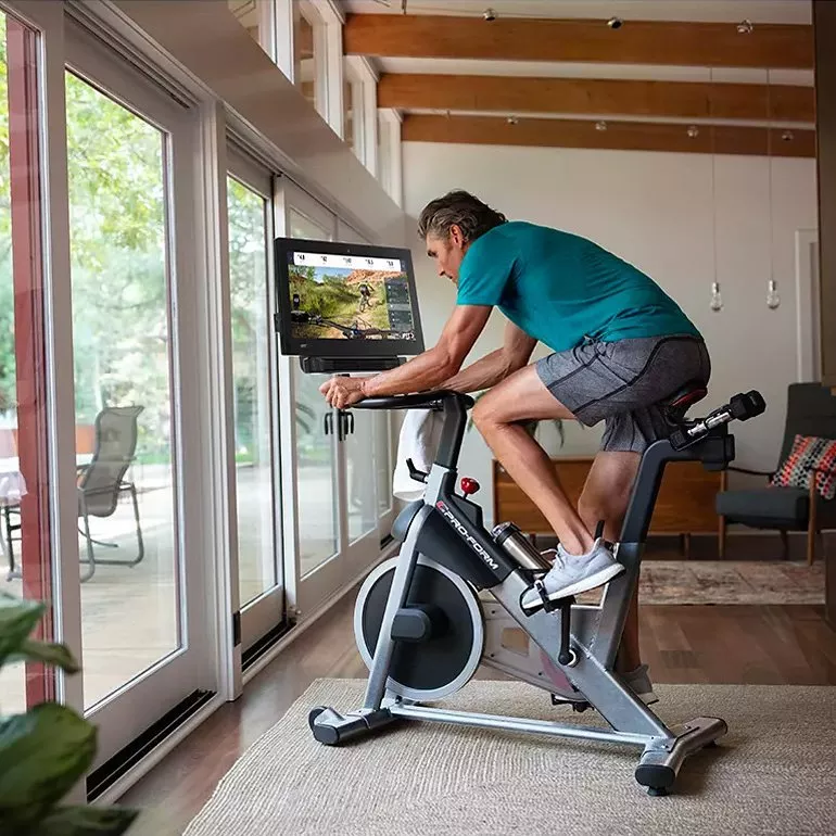 Man cycling on indoor exercise bike with touchscreen training app, in bright modern living room by windows