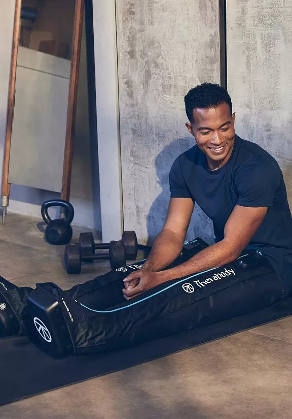 Man using Therabody leg compression boots on gym mat, smiling during recovery with kettlebell and weights nearby