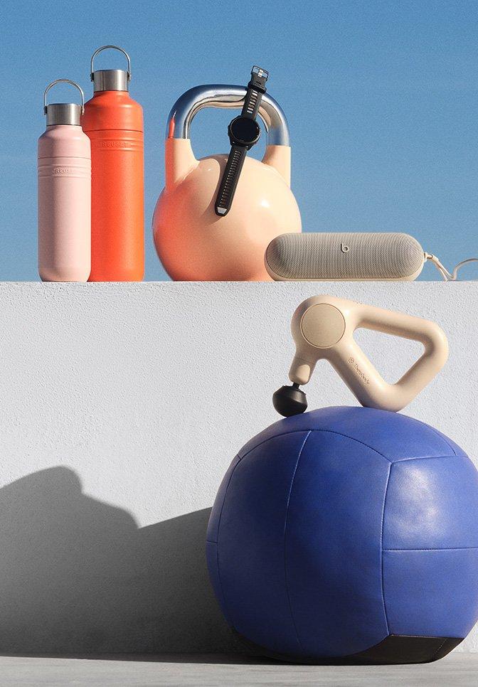 Fitness gear including two water bottles, a peach kettlebell with a smartwatch, a Beats speaker, a massage gun, and a blue medicine ball, all displayed against a minimal outdoor backdrop.
