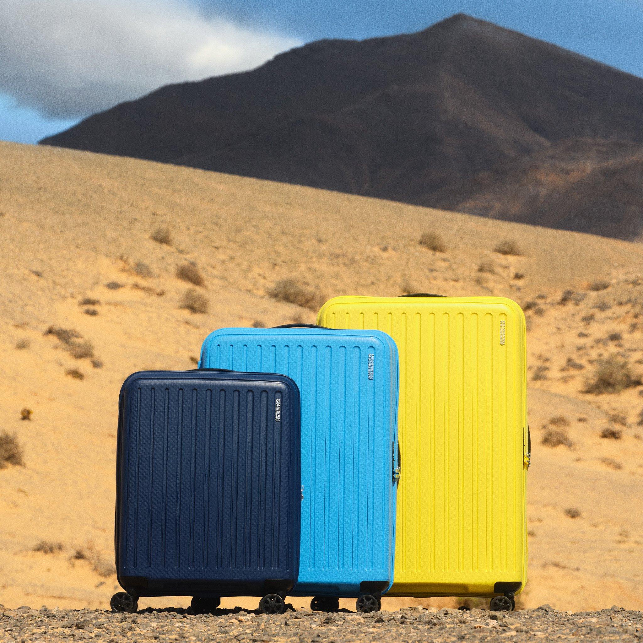 Three hard-shell suitcases in navy blue, sky blue, and yellow are placed in size order on rocky desert ground with sandy hills and a mountain in the background.