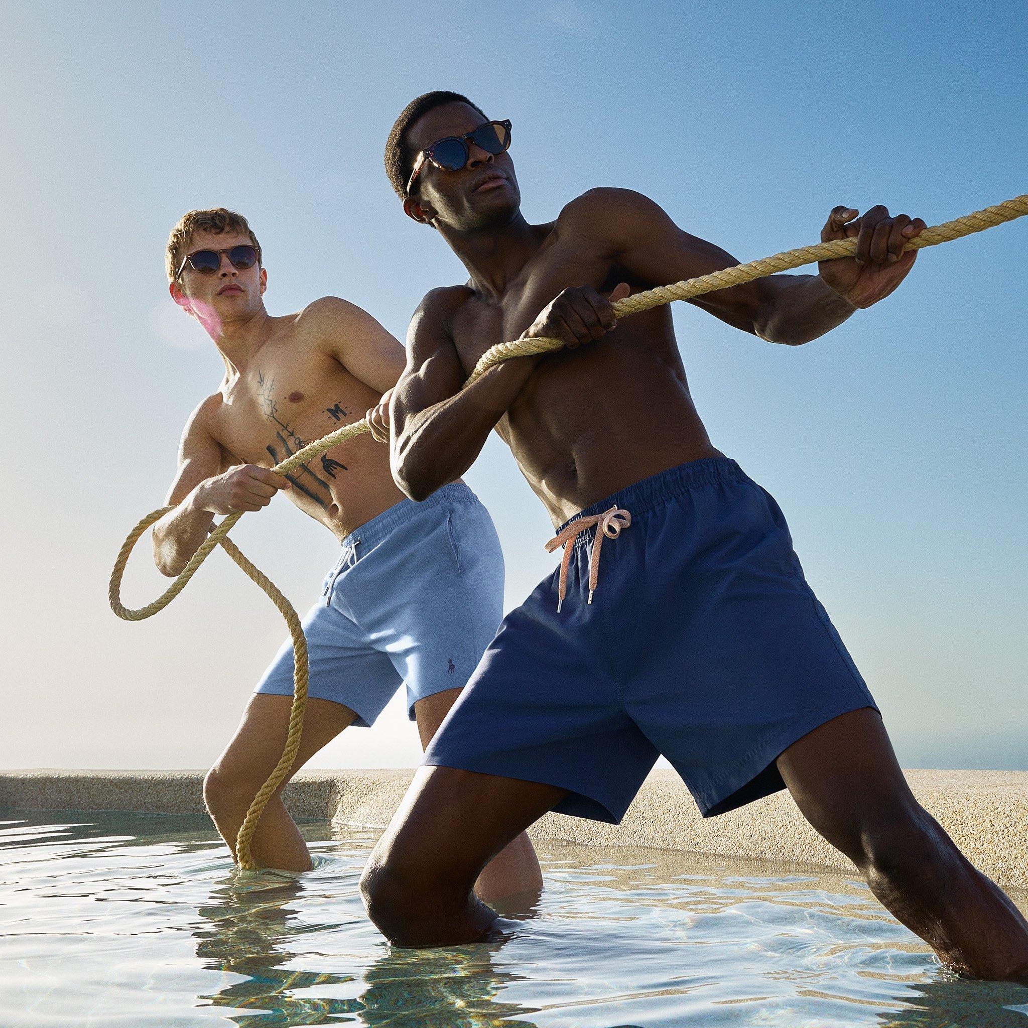 Two men in swim shorts and sunglasses playfully pull a rope in shallow water under clear blue skies.
