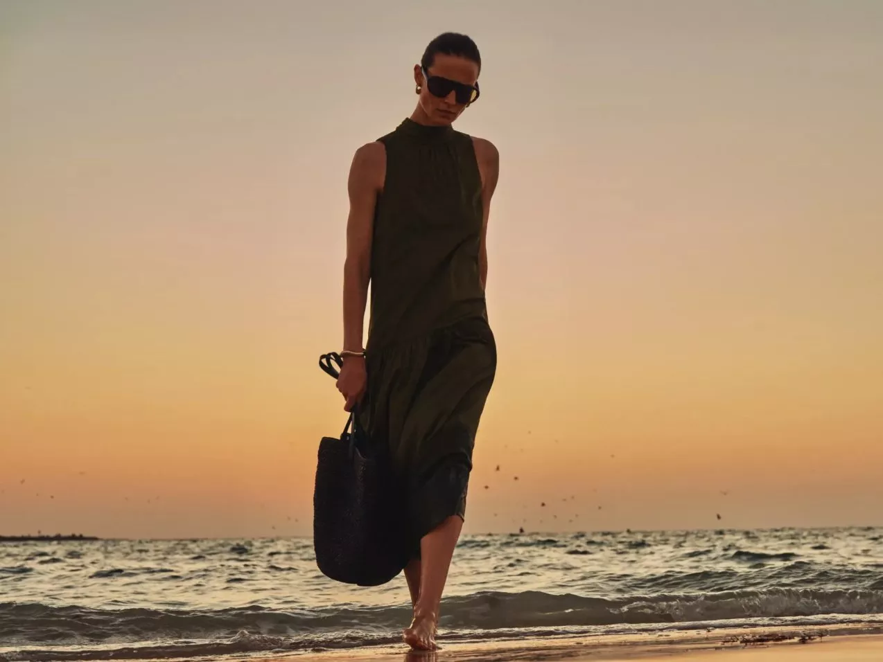 Woman in dark green sleeveless dress walking barefoot on the beach at sunset, holding a bag
