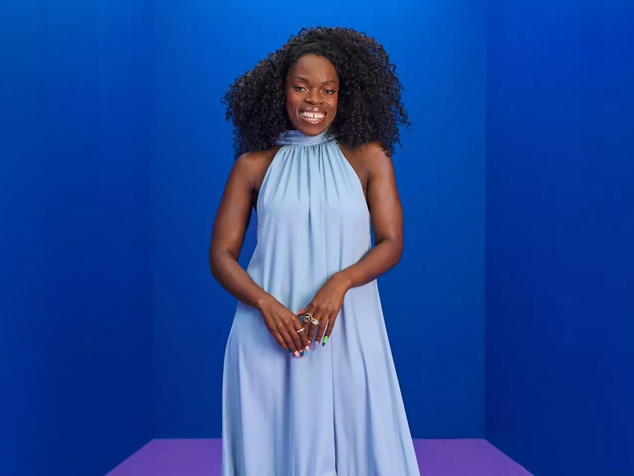 Nana Acheampong in a light blue halter maxi dress with curly hair, standing in a bright blue studio backdrop