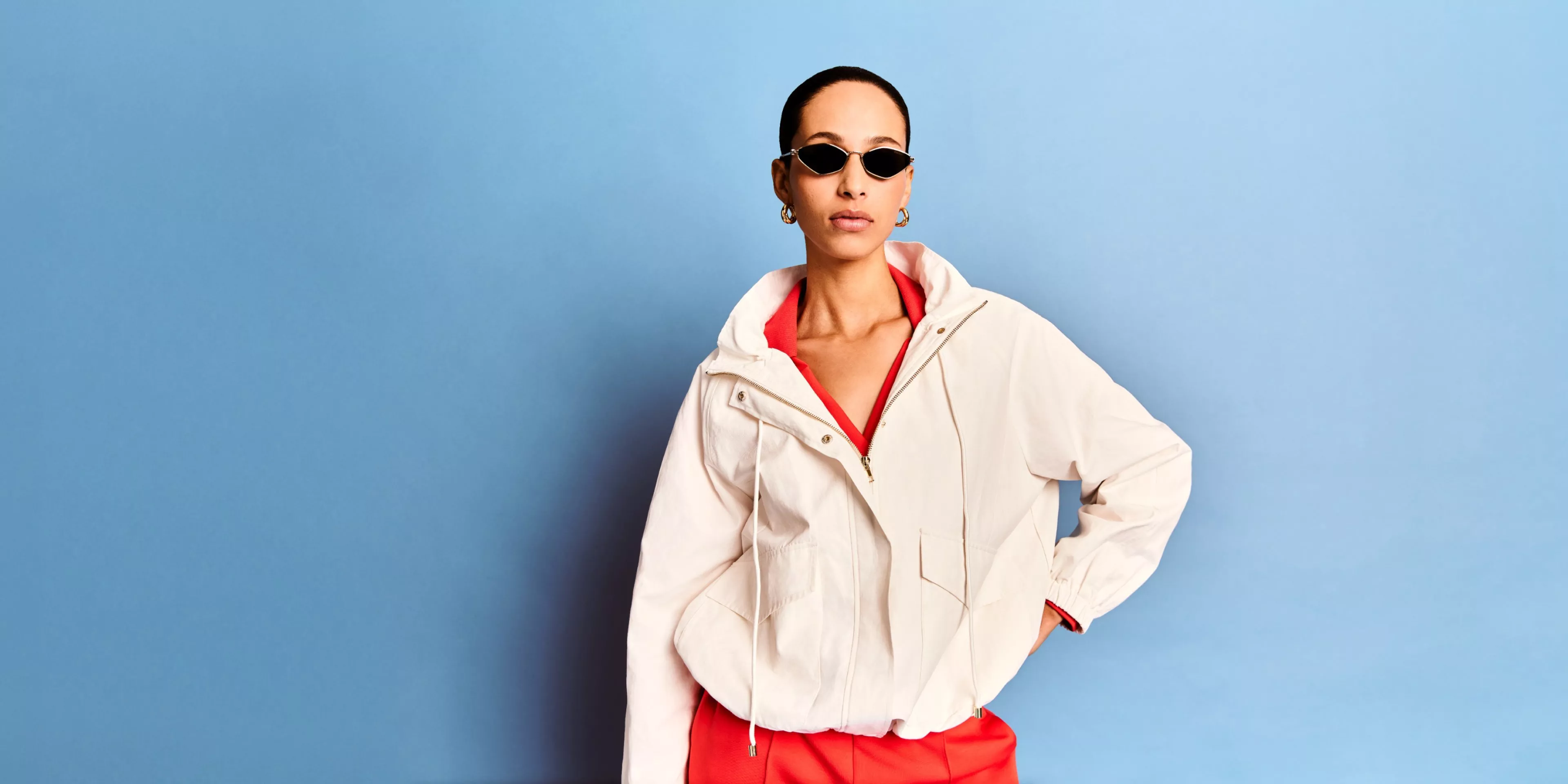 Woman in white jacket and red outfit wearing sunglasses, posing against a blue studio backdrop
