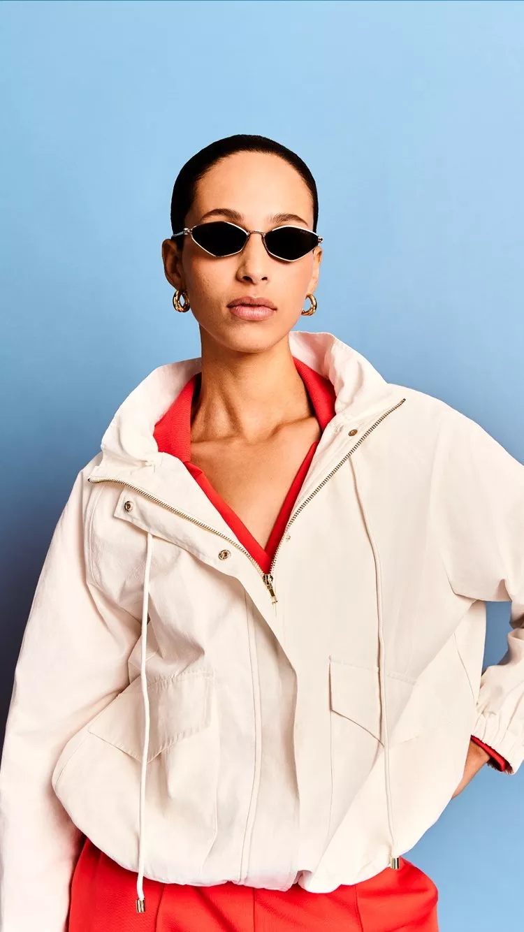 Woman in white jacket and red outfit wearing sunglasses, posing against a blue studio backdrop