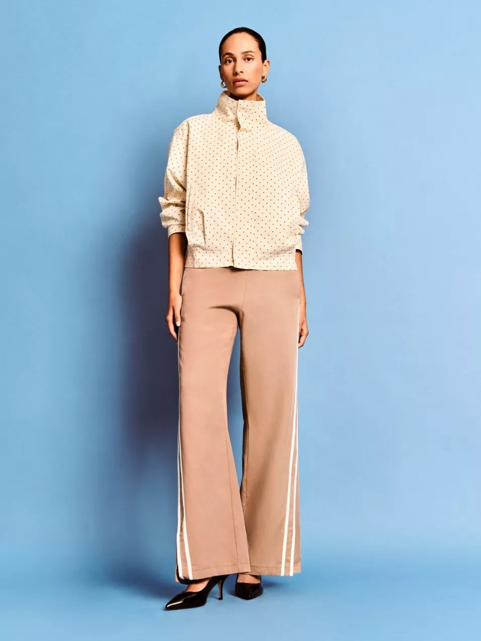 A woman in a beige high-neck jacket and wide-leg trousers on a blue backdrop