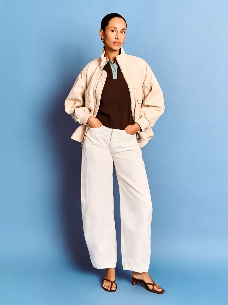 Woman modelling beige jacket with brown polo, white wide-leg trousers and sandals on blue studio backdrop