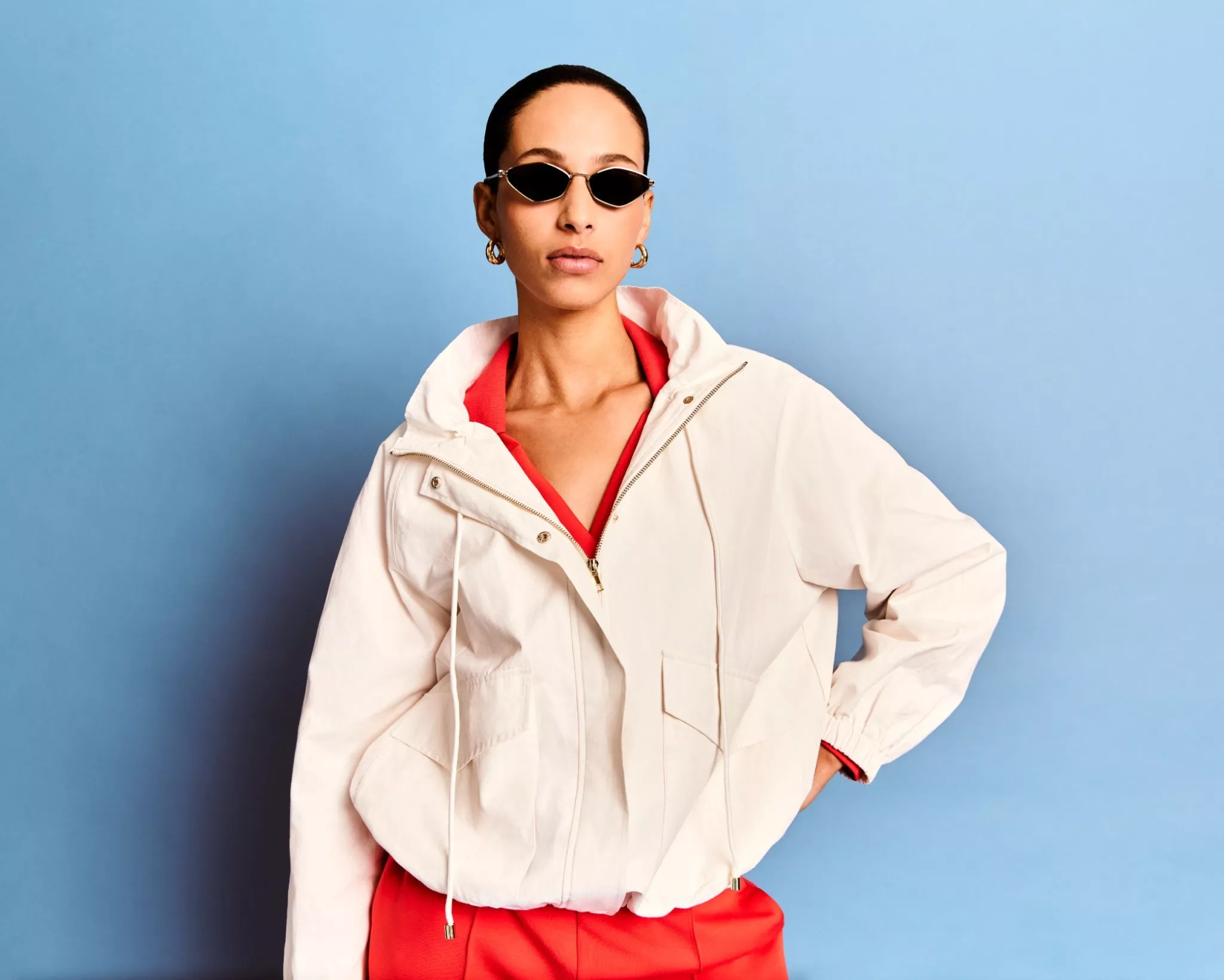 Woman wearing black sunglasses and white jacket over red outfit against a blue studio background