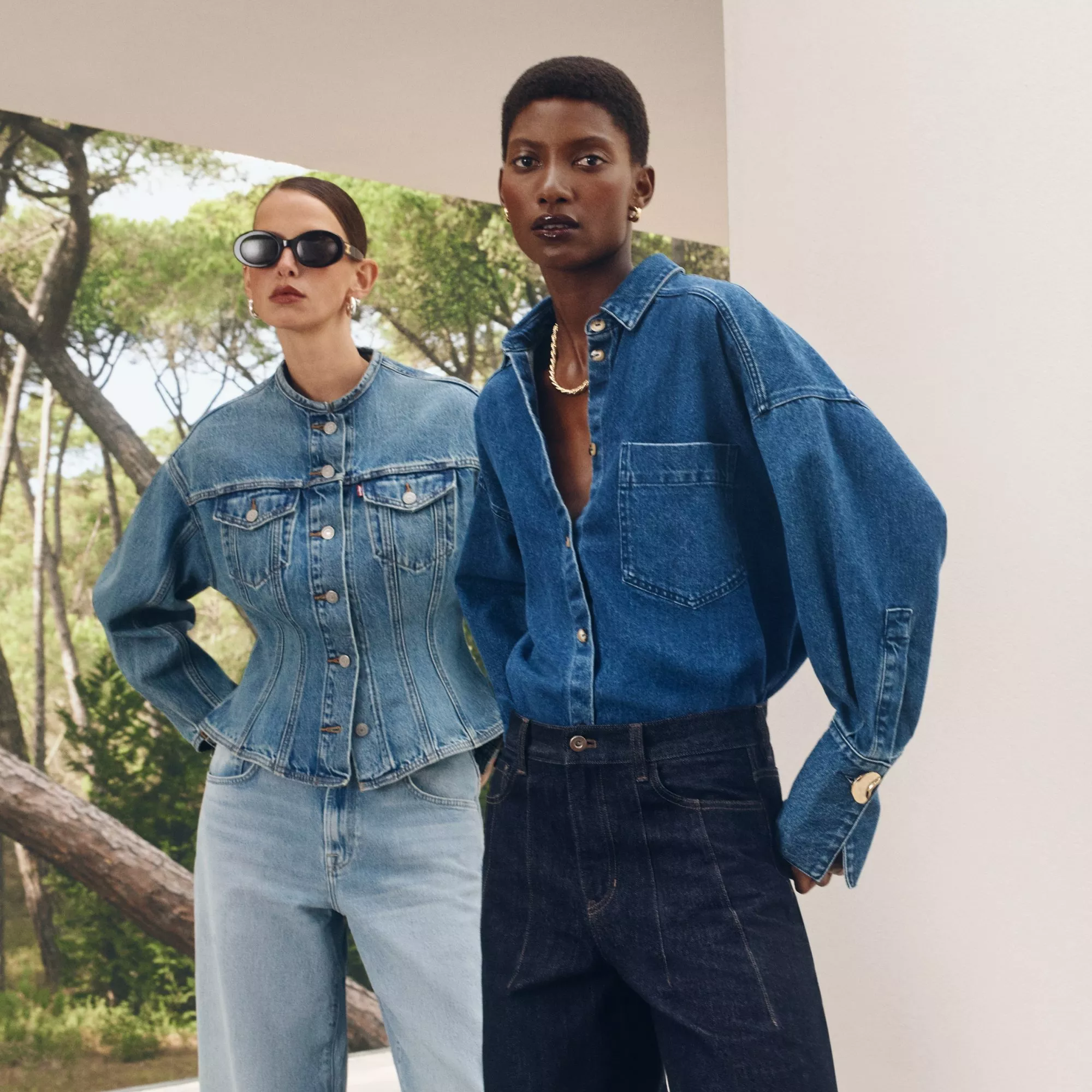 Stylish duo in denim outfits pose outdoors; one in a fitted jacket, the other in a loose shirt. Trees in the background.