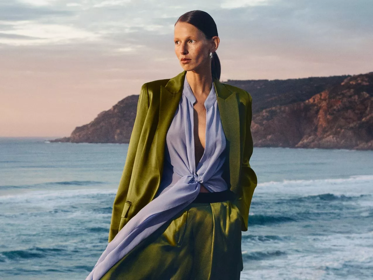 Model in a green blazer and blue blouse stands by the sea at sunset, with cliffs in the background.