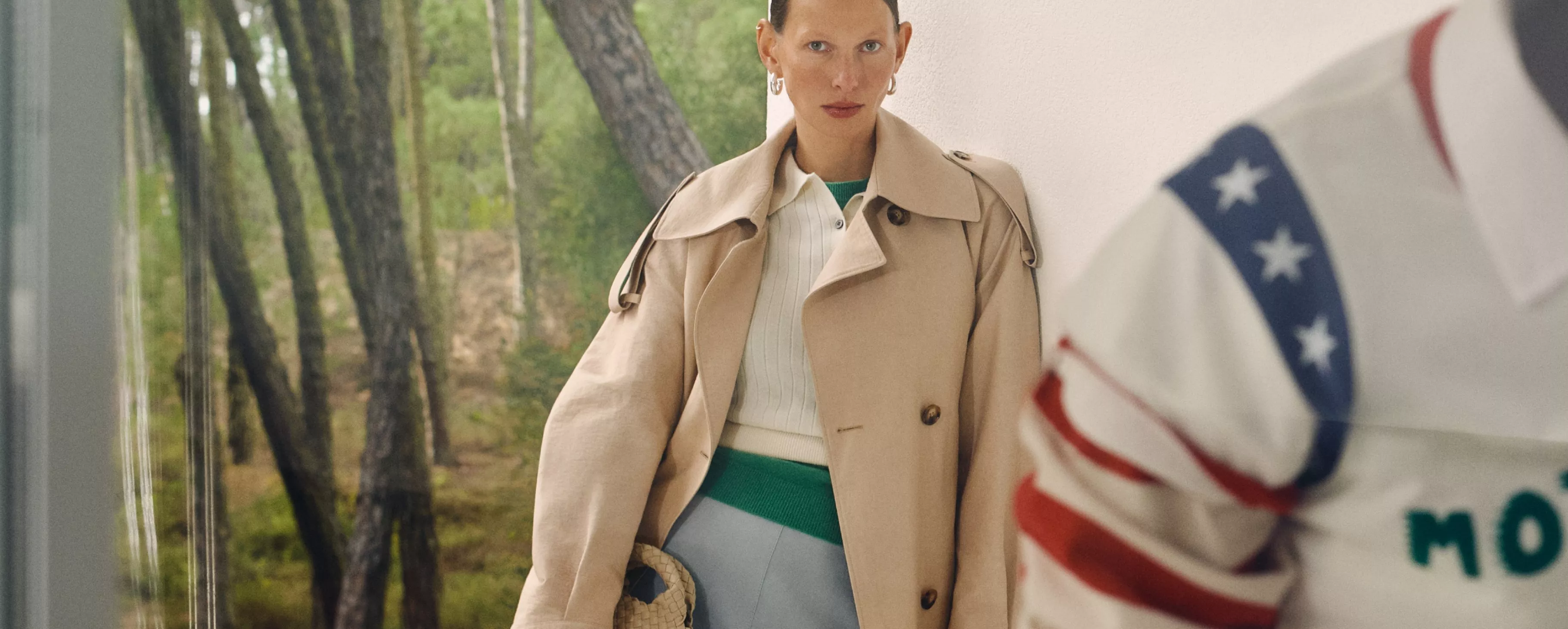 Woman in a beige trench coat with a green and white top, standing indoors with a forest view behind her.