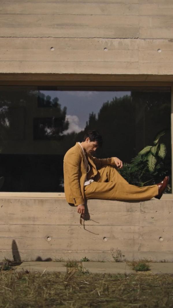Man in a mustard suit sits on a ledge by a modern building with reflective glass, holding sunglasses.