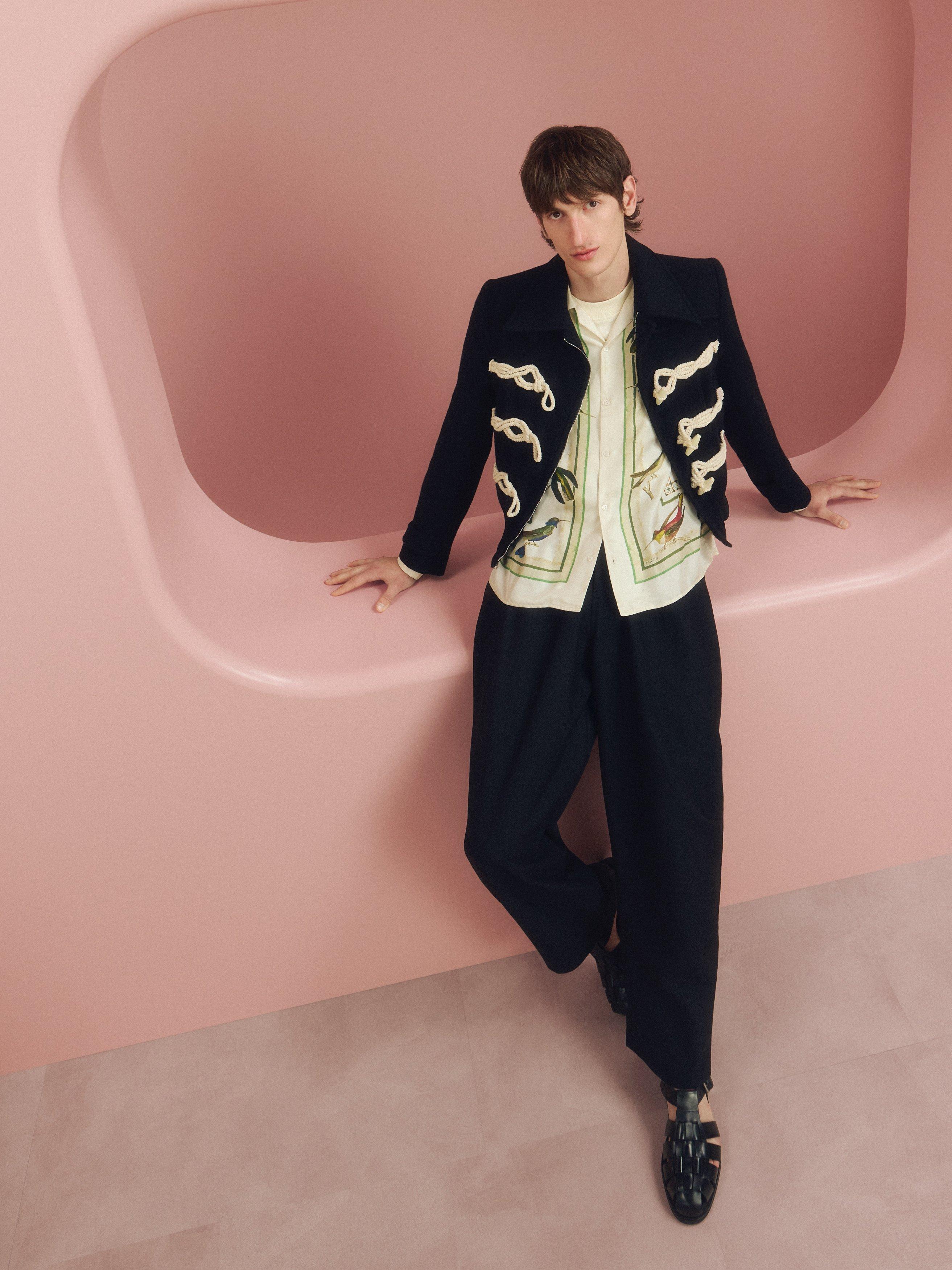 Image of a male model leaning on a pink backdrop wearing a black military jacket and trousers