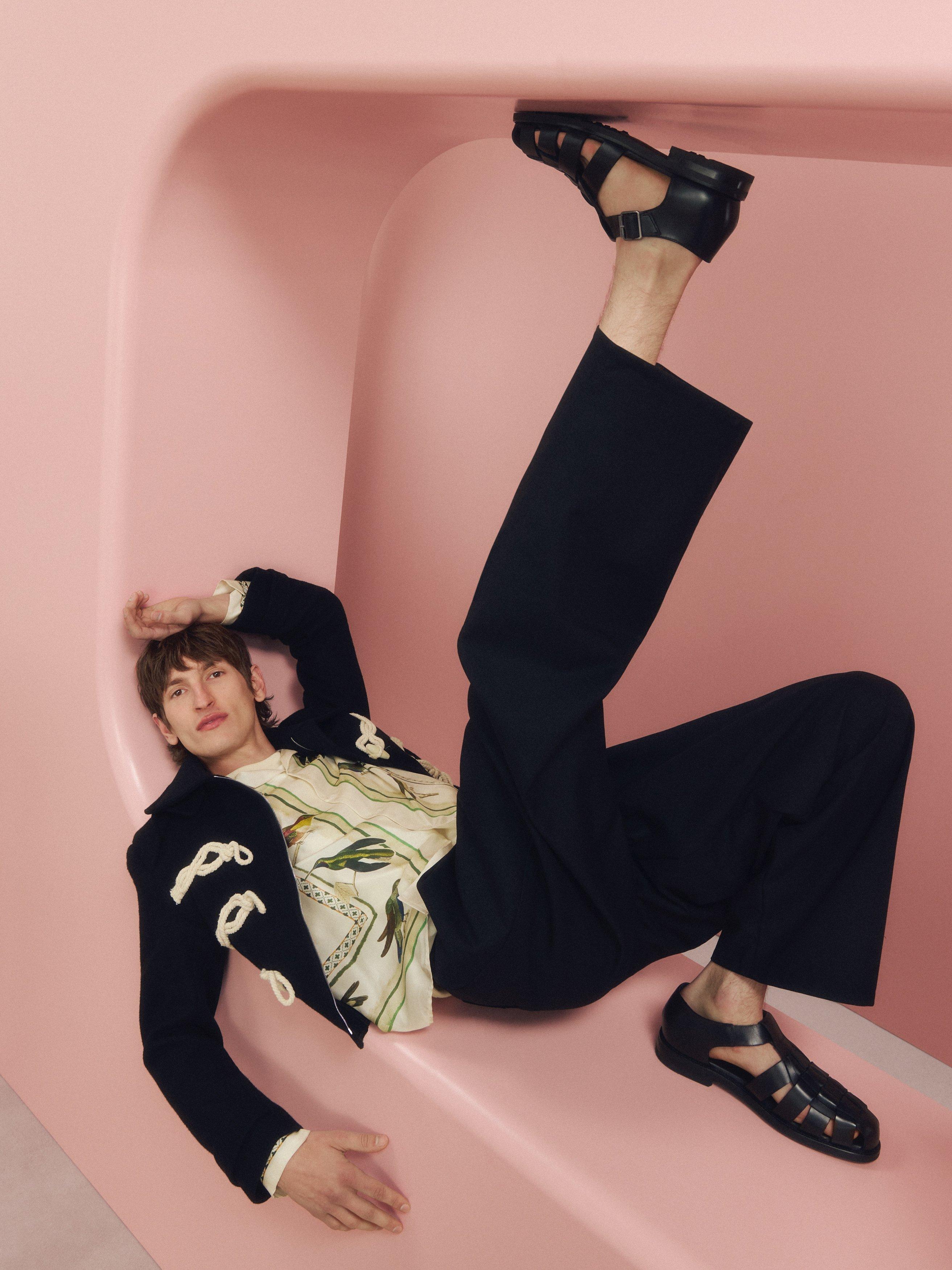 A male model lying on a pink built backdrop wearing a military jacket