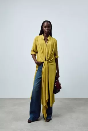 Studio fashion portrait of woman in mustard yellow draped blouse and wide-leg jeans, holding small handbag.