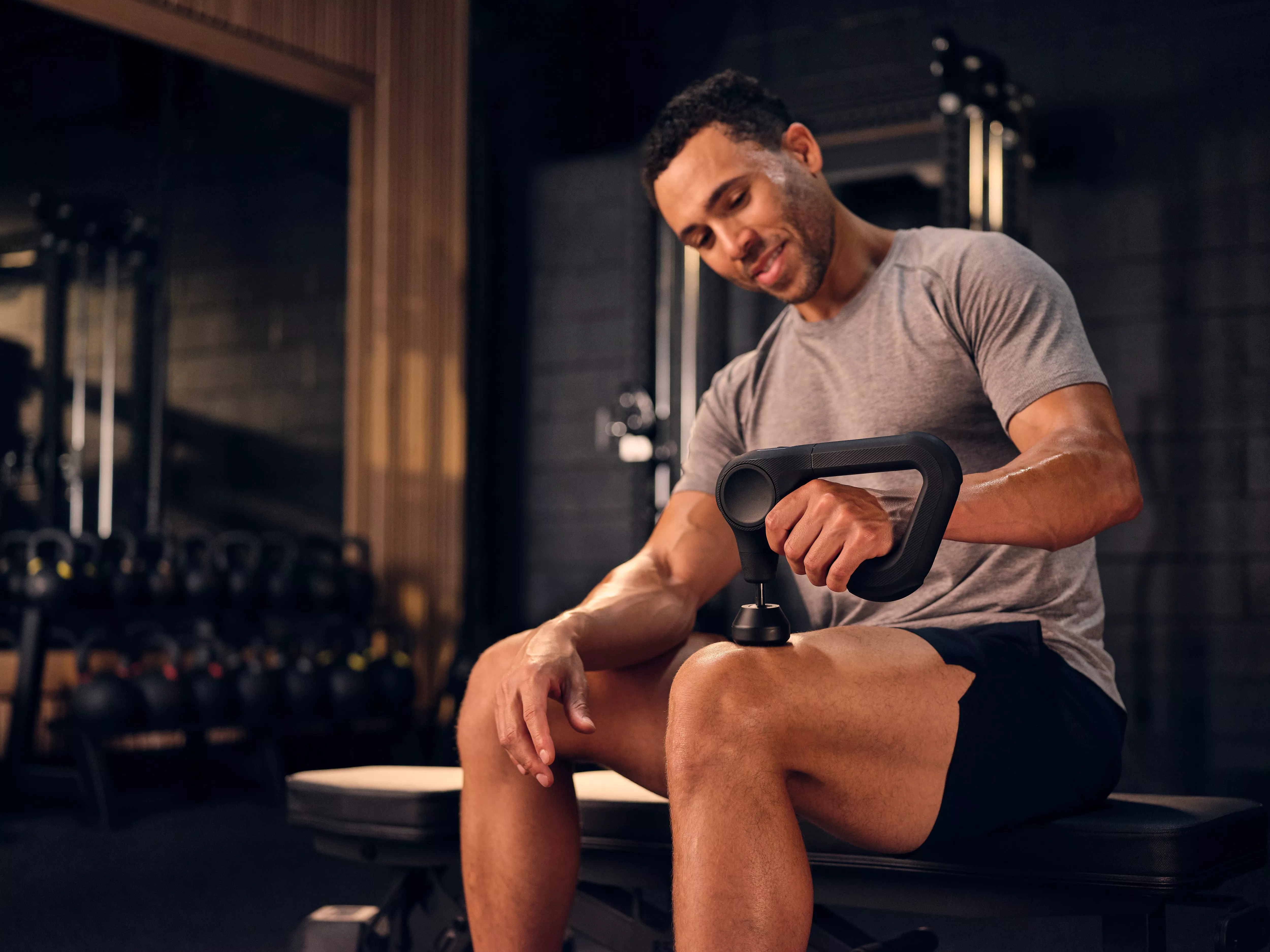Man using handheld percussion massage gun on thigh in gym, post-workout muscle recovery and fitness training.