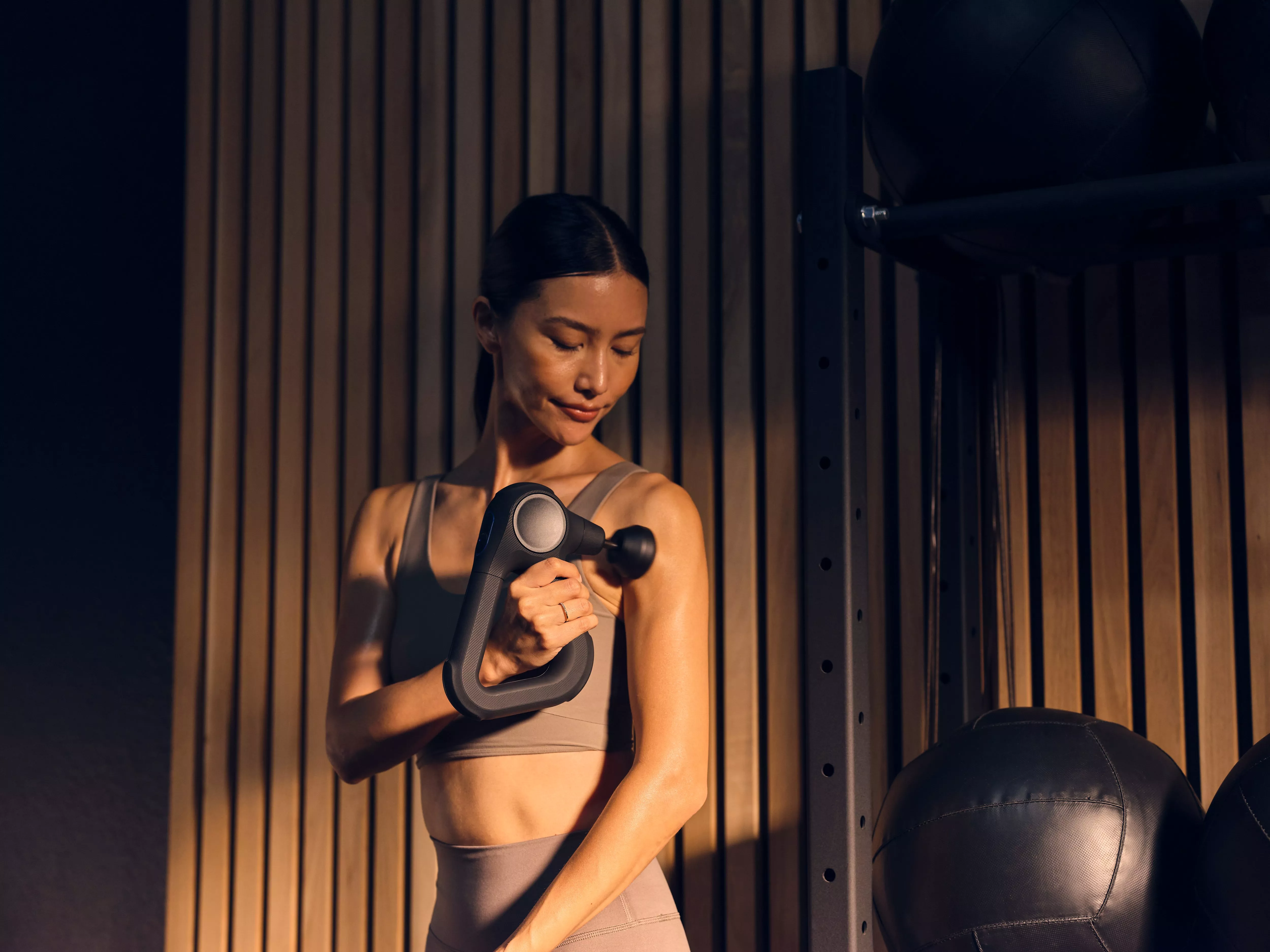 Woman using handheld percussion massage gun for muscle recovery in a modern gym with wooden wall panels