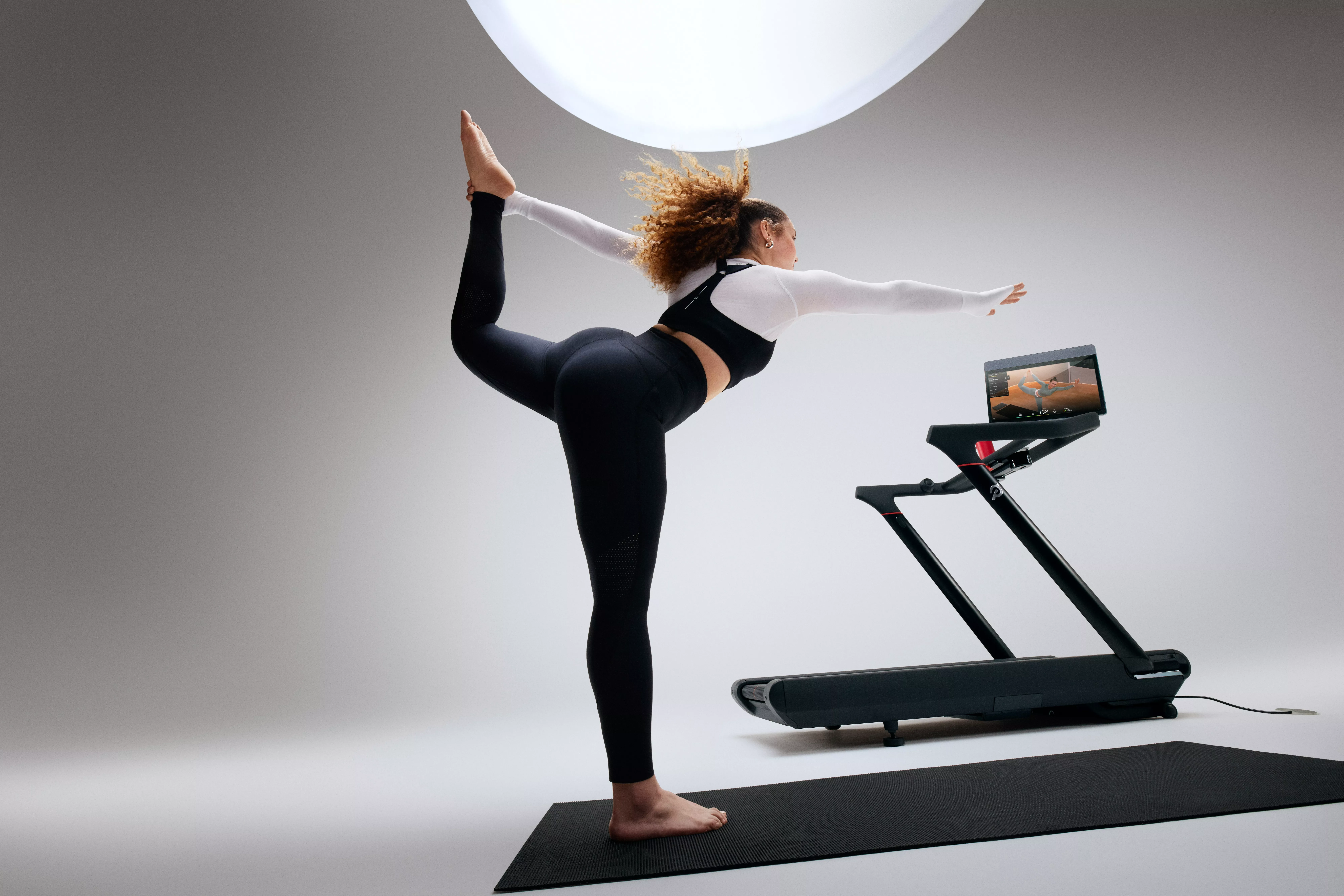 Woman doing dancer pose yoga stretch beside smart treadmill in modern home gym studio with fitness screen