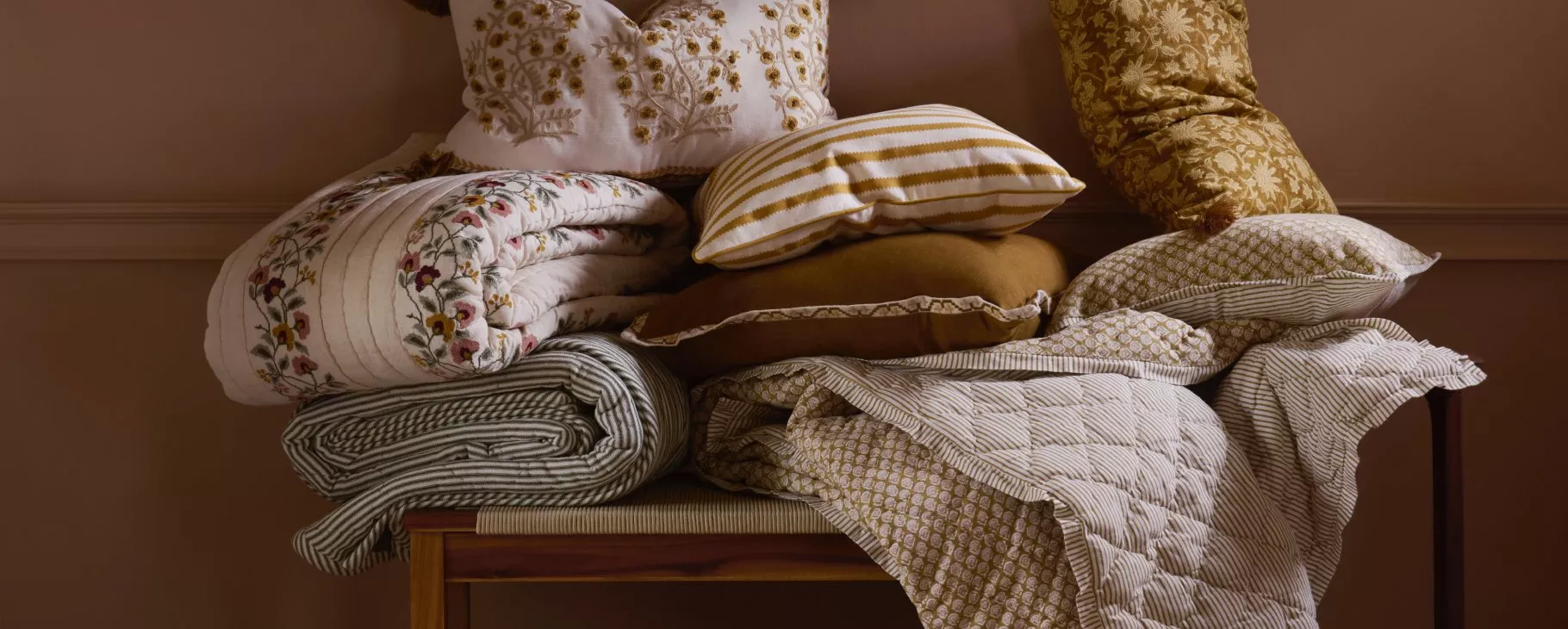 Stacked decorative cushions and folded quilts on a bench, cosy neutral bedroom textiles with floral embroidery