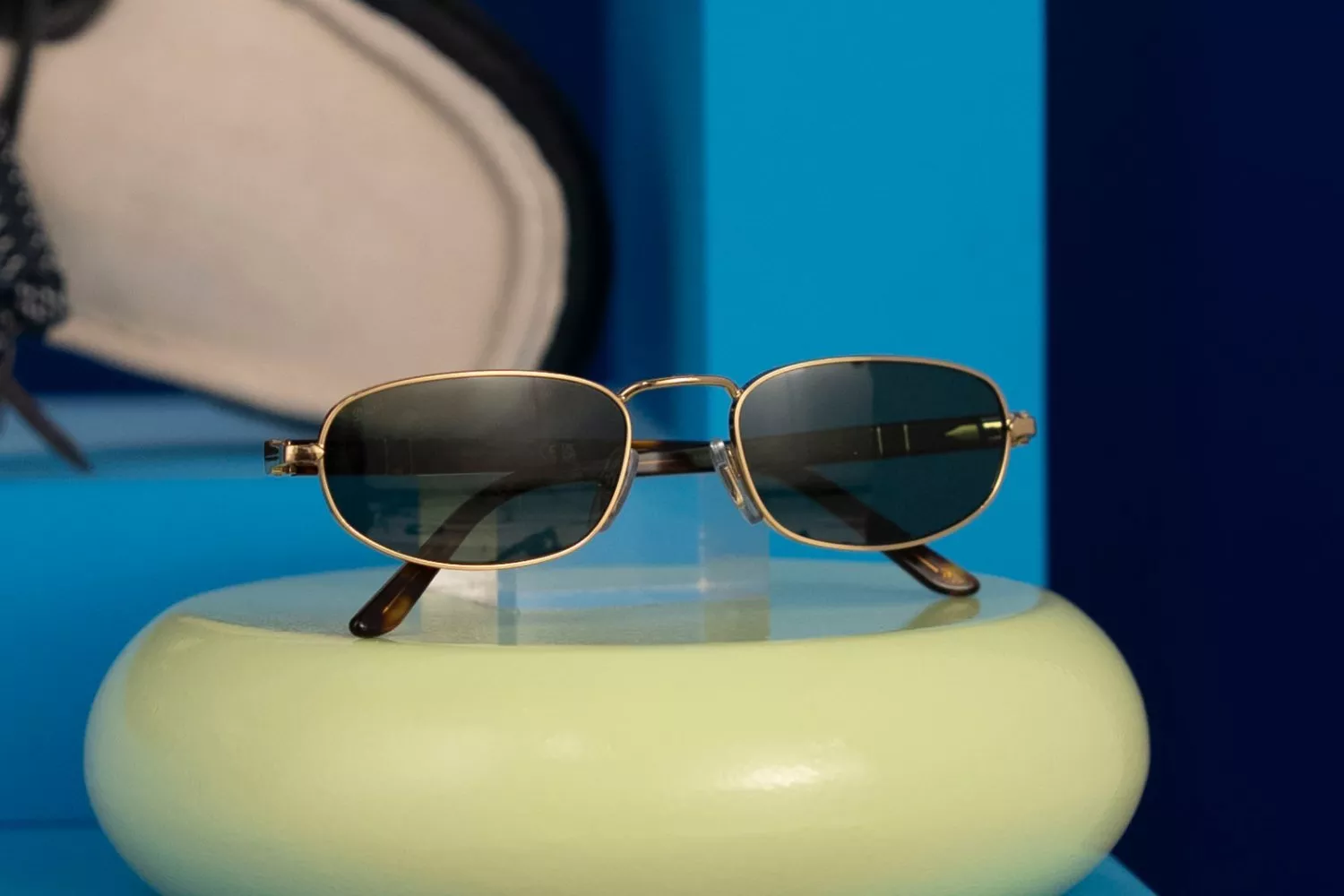 Gold metal Persol oval sunglasses with dark lenses on a green stand, set against a blue display background