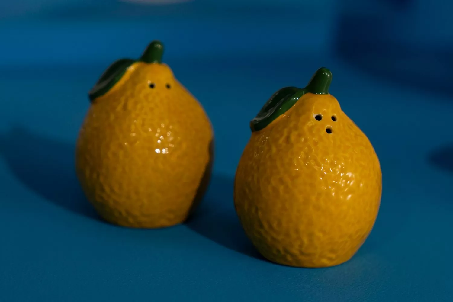 Lemon-shaped ceramic salt and pepper shakers on a blue surface, bright yellow with green stems and leaves