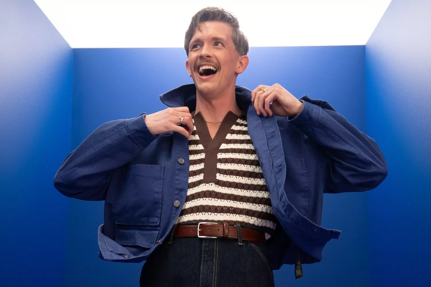 Smiling Stephen Libby adjusting blue jacket over striped knit top in a blue studio, modern menswear fashion portrait