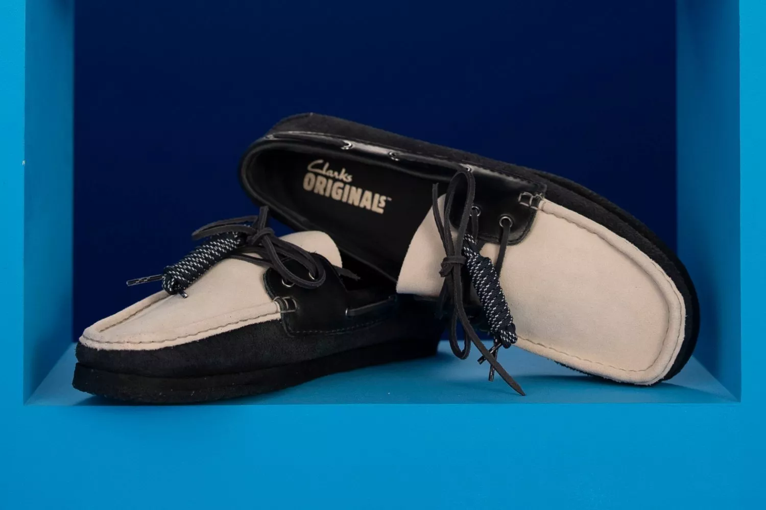 Pair of Clarks Originals boat shoes in black and cream suede, styled on a blue display background