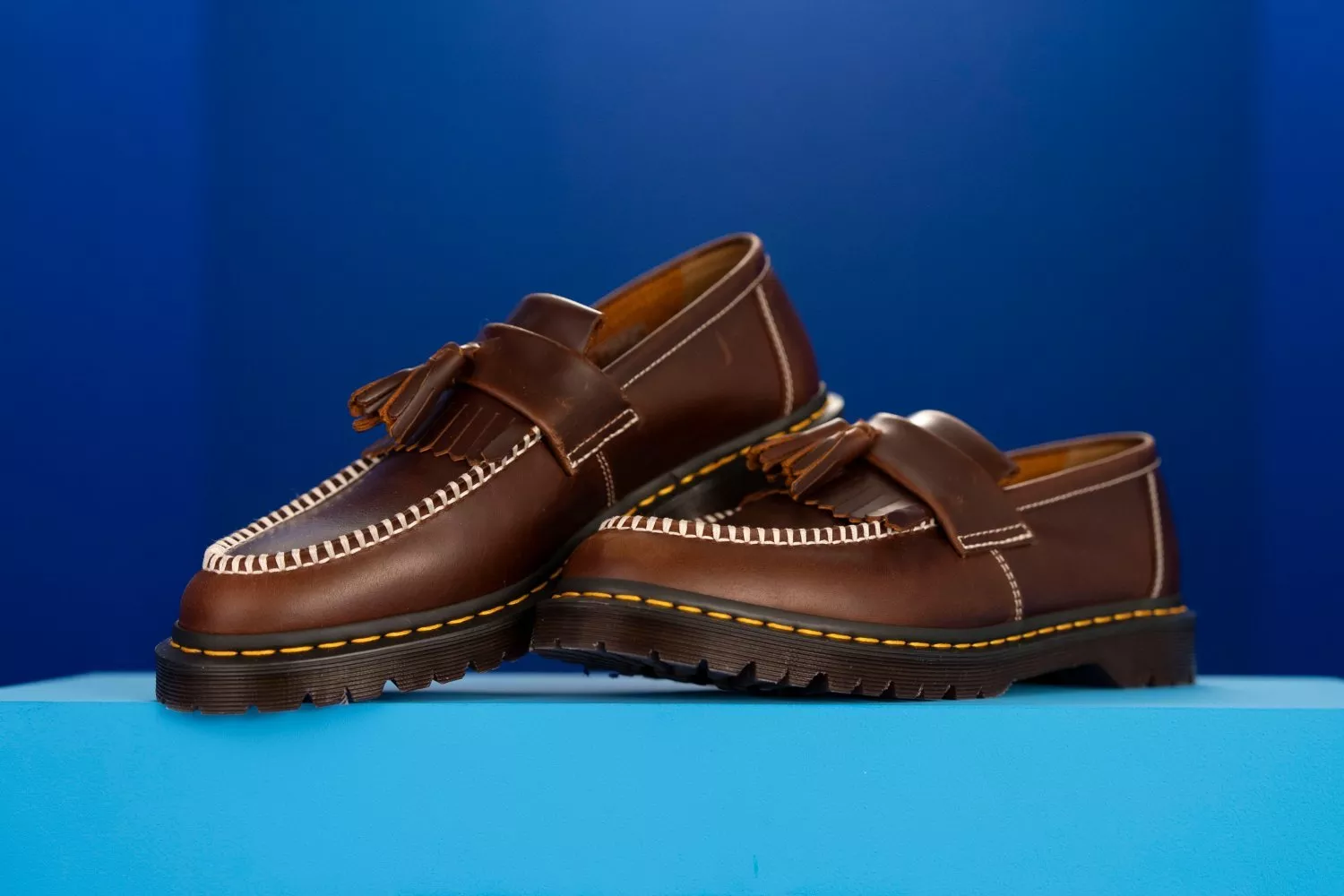 Pair of brown leather tassel loafers with chunky soles and contrast stitching on a blue studio background