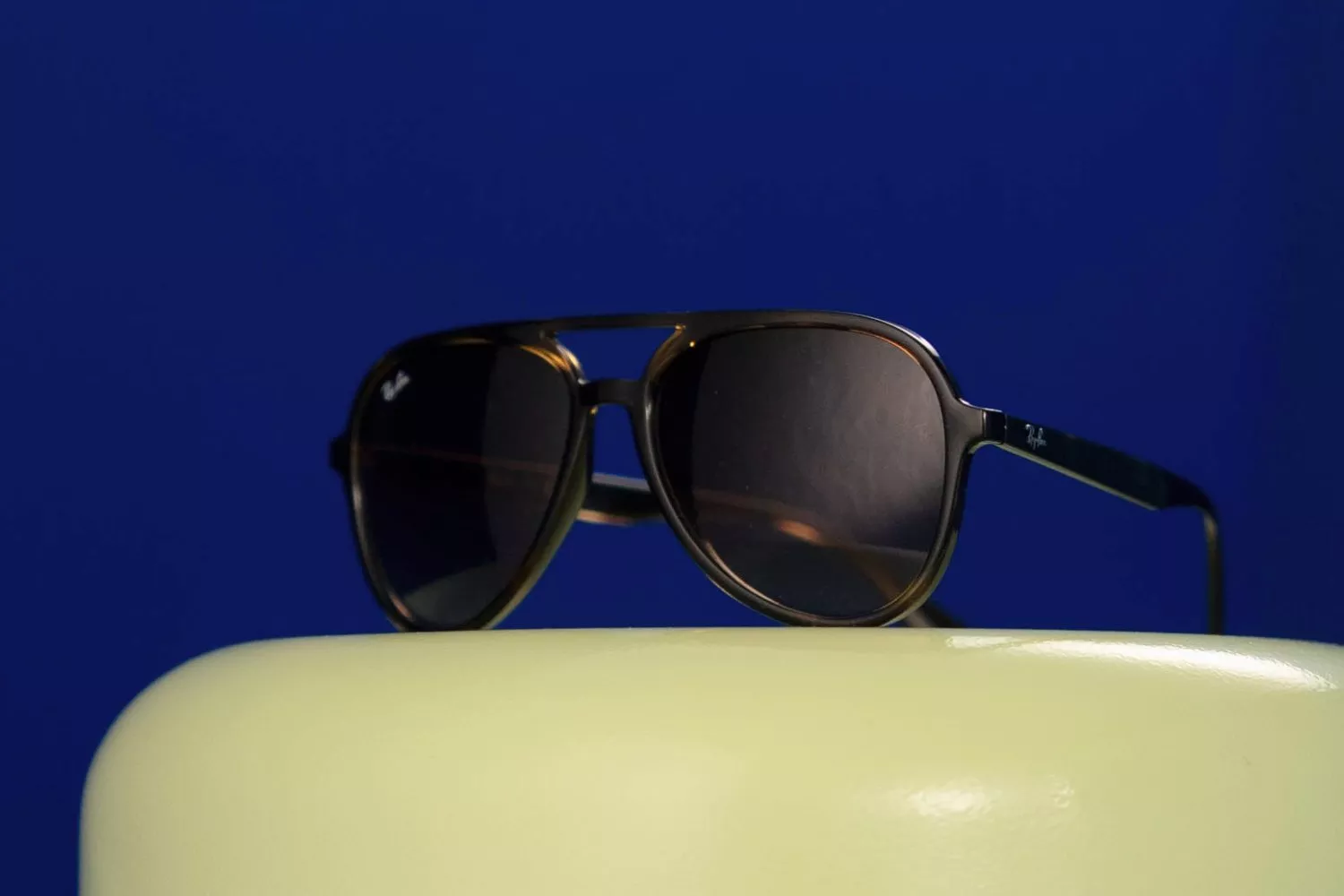 Black aviator sunglasses on a green plinth against a deep blue backdrop, stylish eyewear product photo