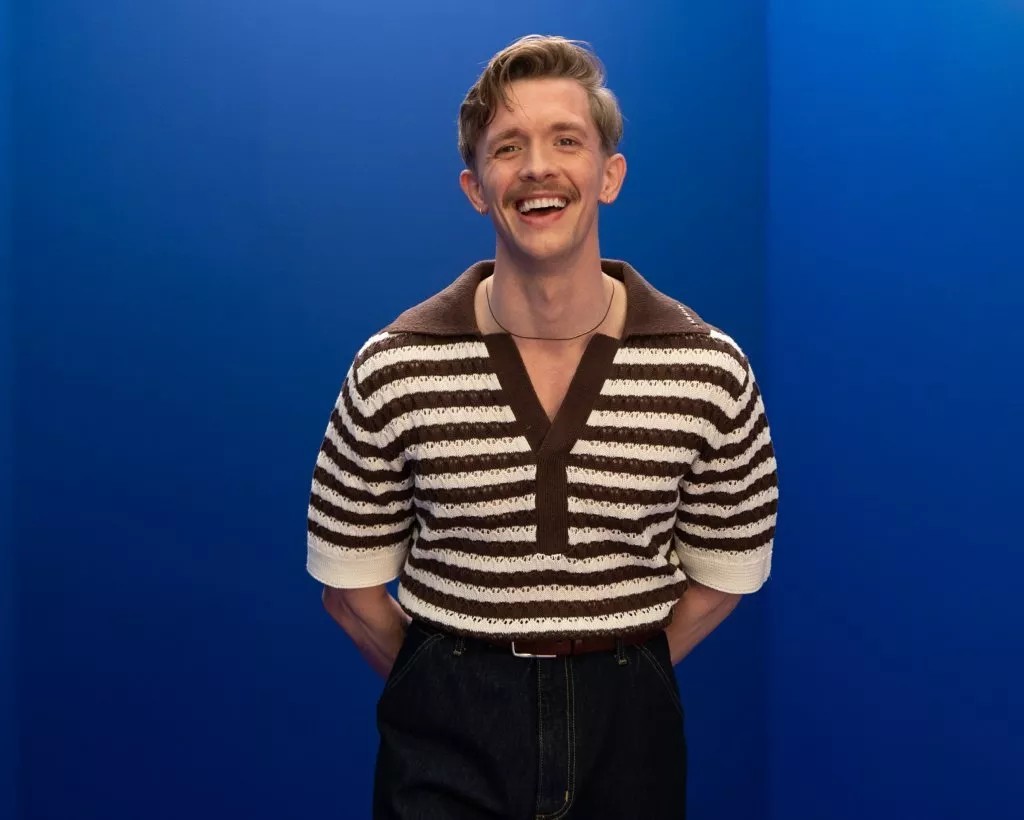 Smiling man in striped knit polo and dark trousers, studio portrait on bright blue background, hands behind back