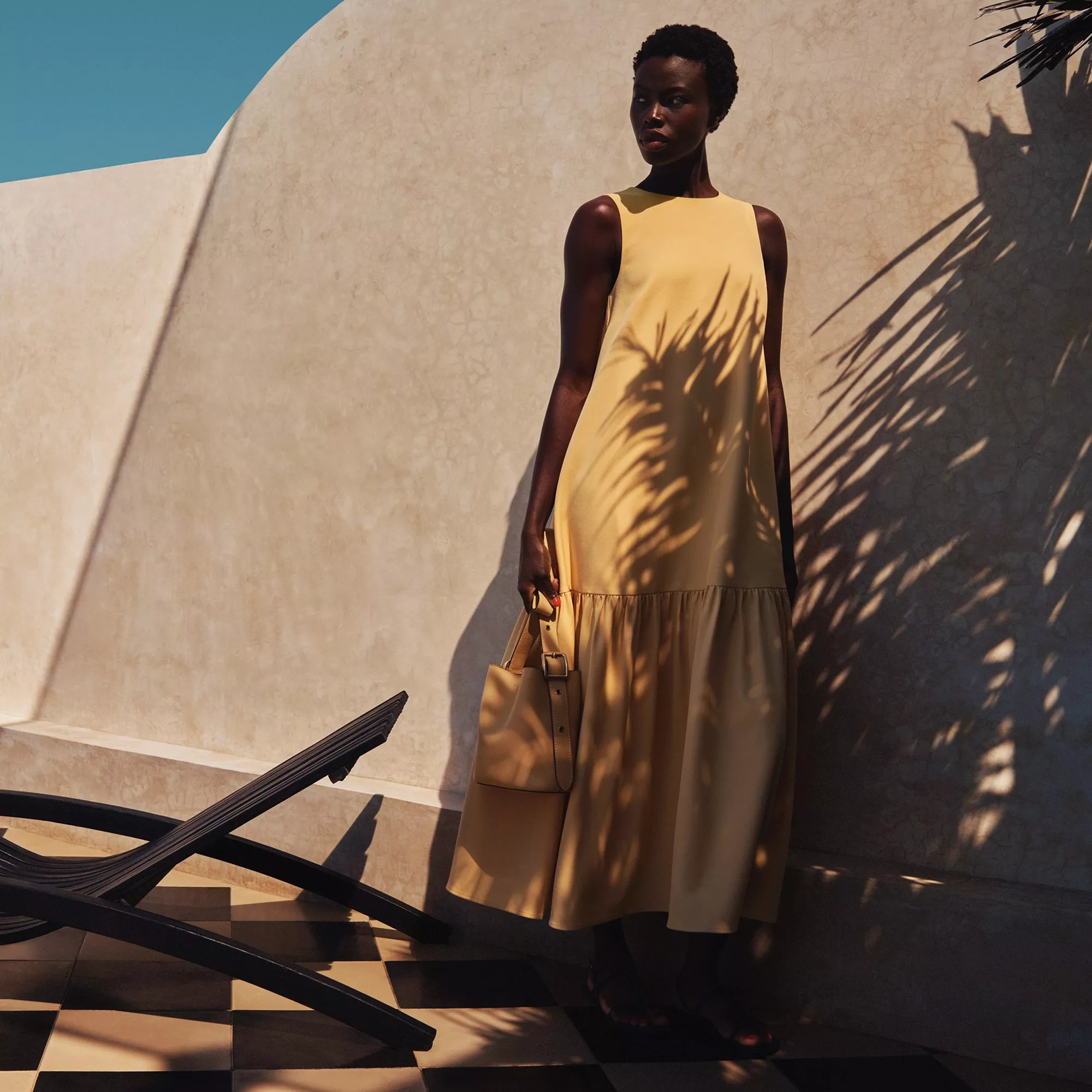 Fashion model in a sleeveless yellow maxi dress holding a tan handbag beside a sunlit stucco wall outdoors
