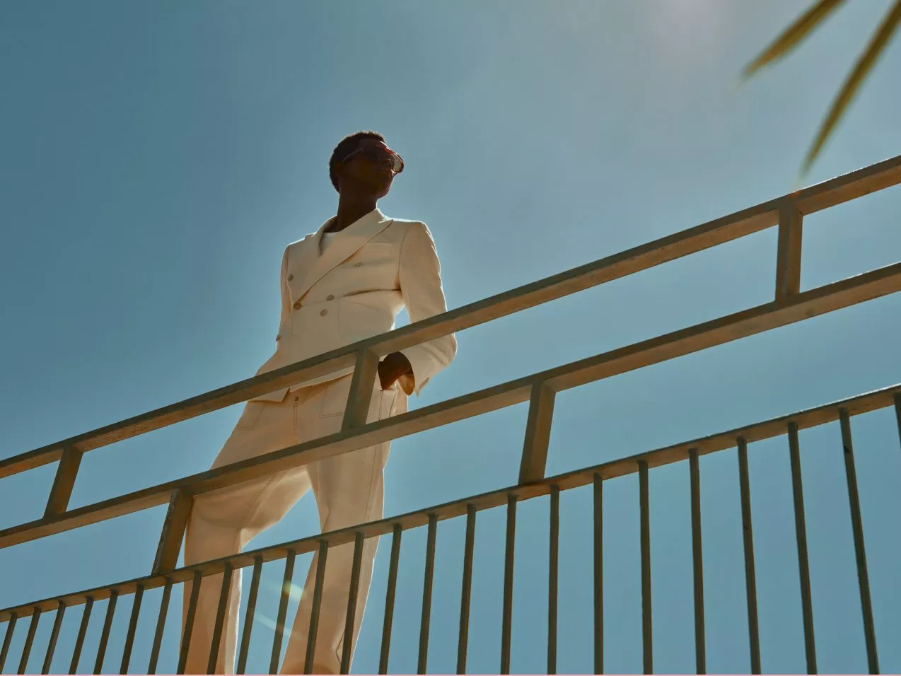 Fashion model in cream double-breasted suit on modern balcony railing against clear blue sky, sunlit style shot