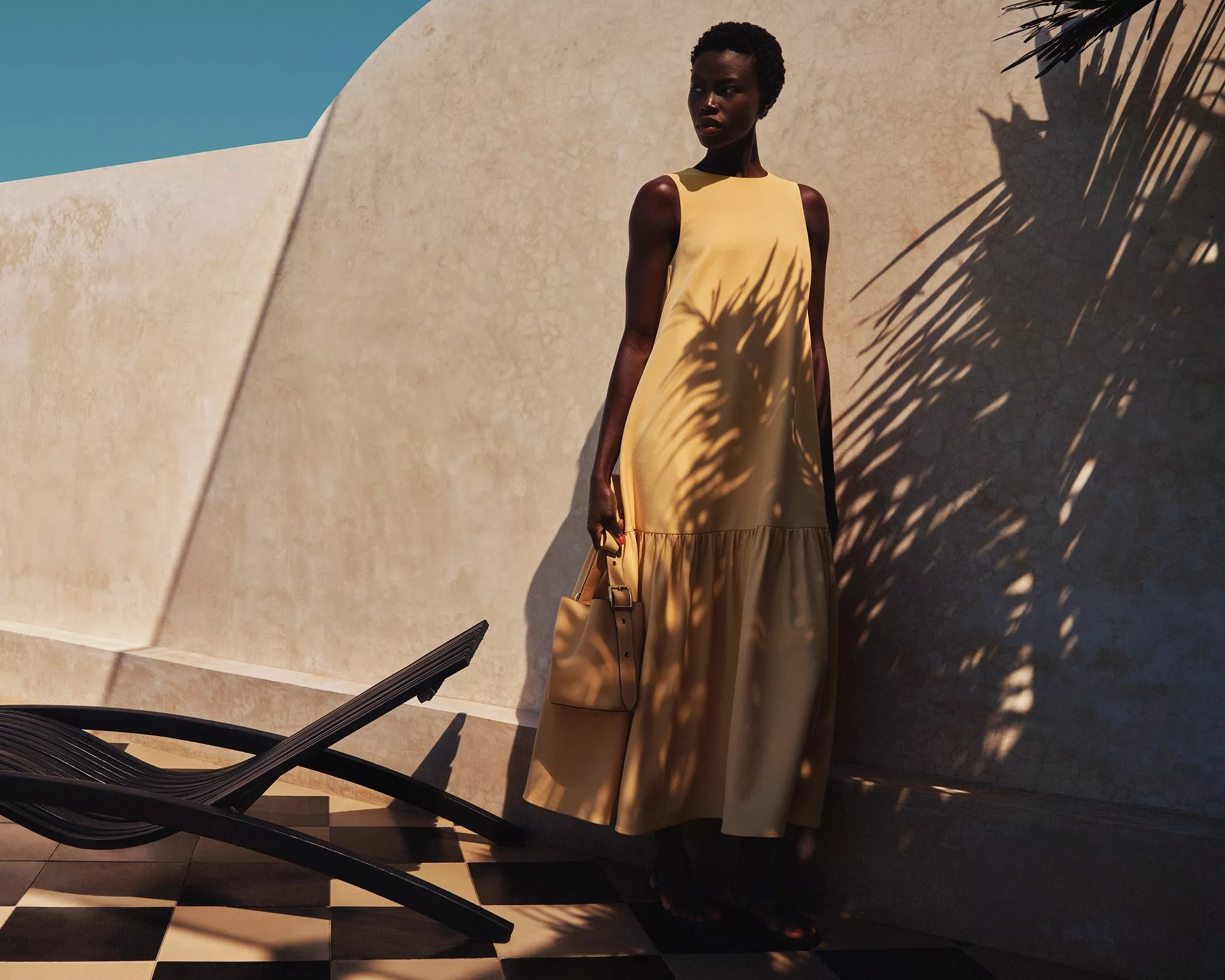Fashion model in a flowing yellow maxi dress holding a tote bag on a sunny terrace with palm shadows