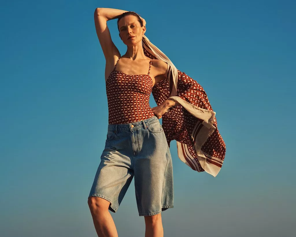 Fashion model in patterned camisole and denim bermuda shorts, holding scarf against blue sky backdrop